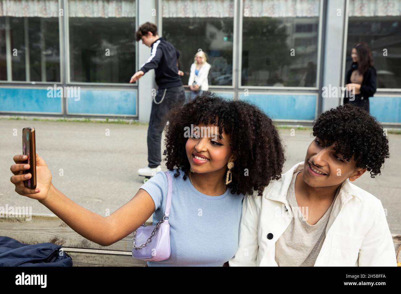 Teenager-Mädchen, das Selfie mit einem männlichen Freund über das Smartphone nimmt, während sie auf der Bank sitzt Stockfoto