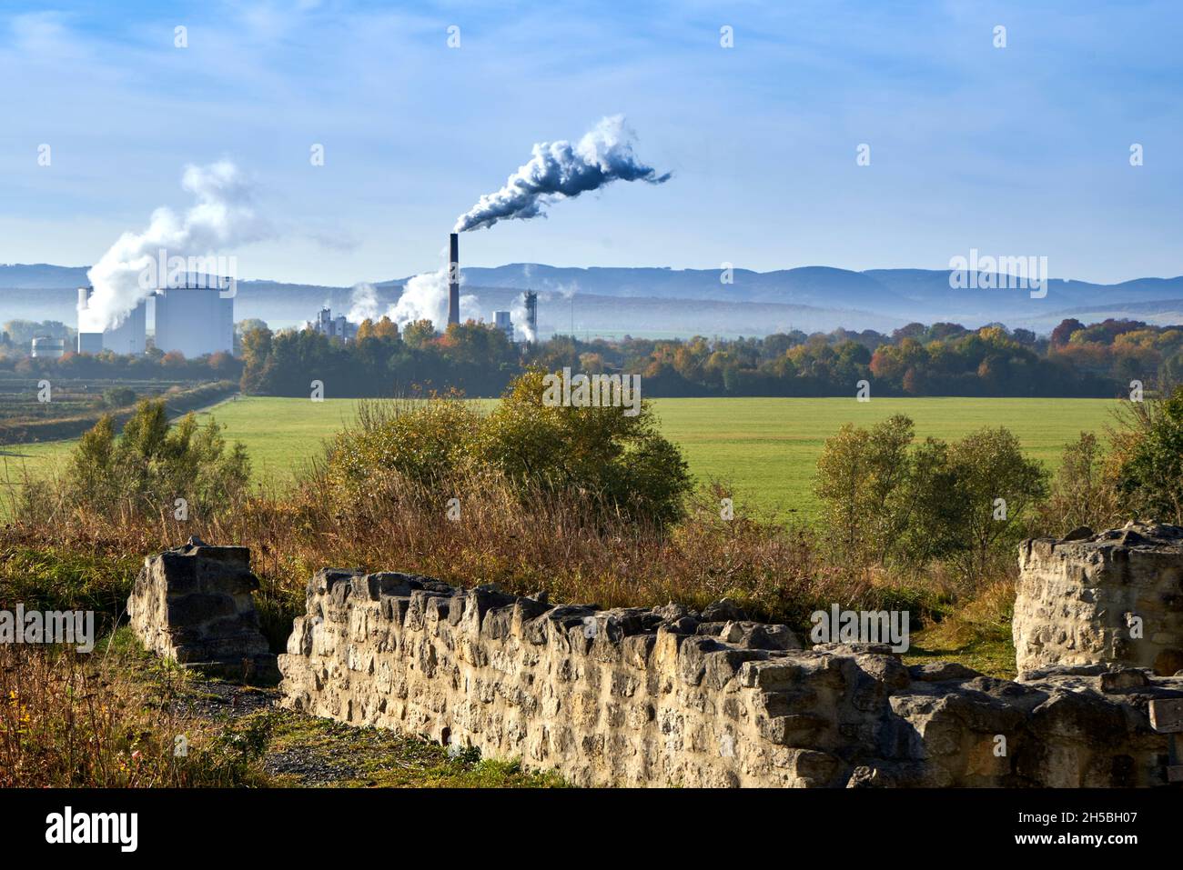 Wände einer alten Ruine aus Naturstein mit einer Fabrik mit rauchenden Kaminen im Hintergrund vor der Gebirgslinie am Horizont Stockfoto