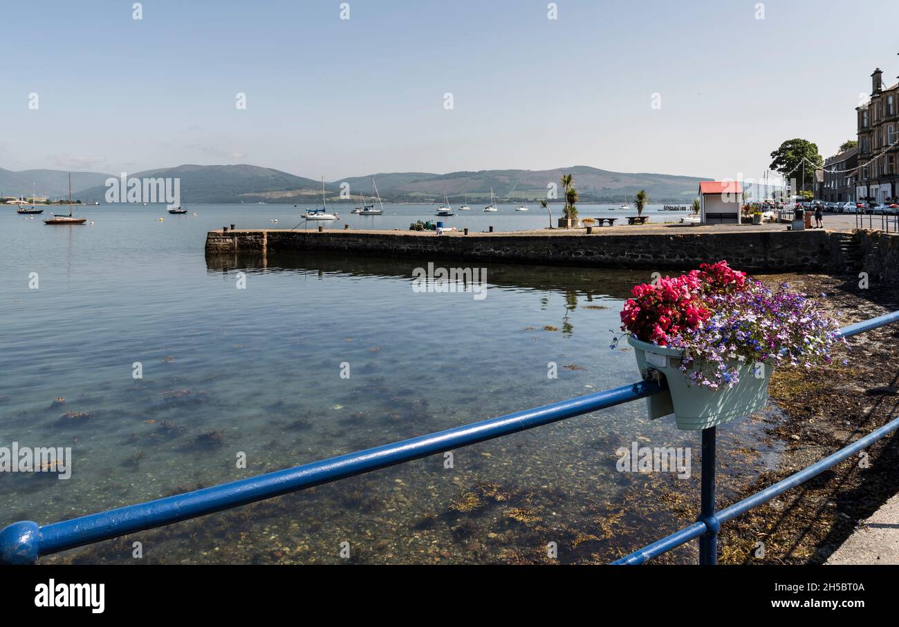 Port Bannatyne Pier und Meeresfront mit Blumen und Palmen, die wie die mediterrane Riviera aussehen, bei strahlendem Sonnenschein mit blauem Himmel, Meer und Yachten. Stockfoto