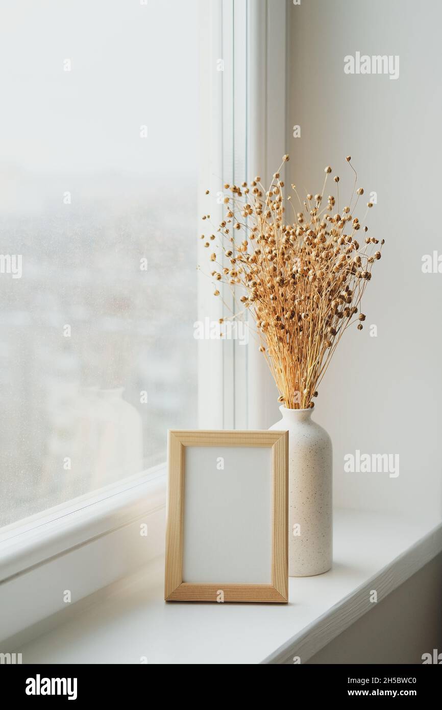 Fotorahmen-Mockup und Vase mit getrockneten Flachsblumen auf Fensterbank. Die Inneneinrichtung des Wohnzimmers im Scandi-Stil. Stockfoto