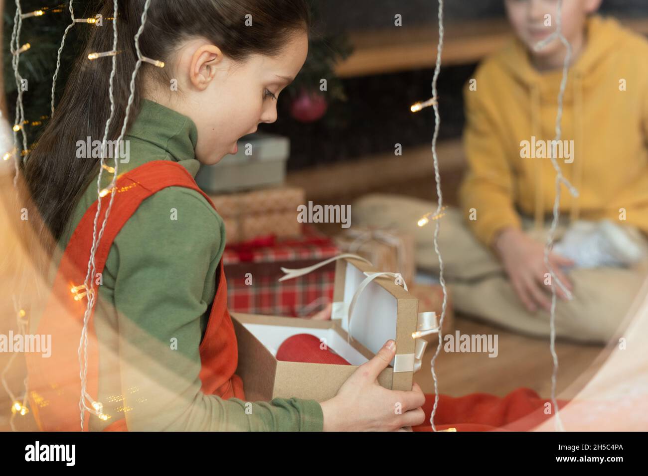 Brunette Mädchen mit offenem Mund sitzt an hängenden Lichtern und Eröffnung Geschenk-Box, sie rempfing unerwartete Weihnachtsgeschenk Stockfoto