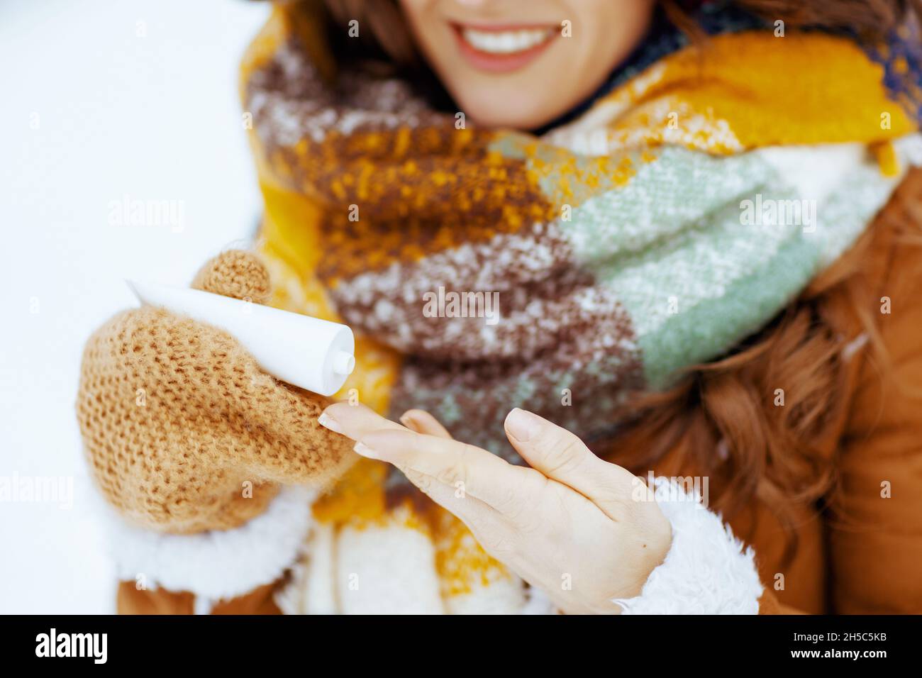 Nahaufnahme einer glücklichen Frau in braunem Hut und Schal mit Fäustlingen mit Handcreme draußen im Stadtpark im Winter. Stockfoto