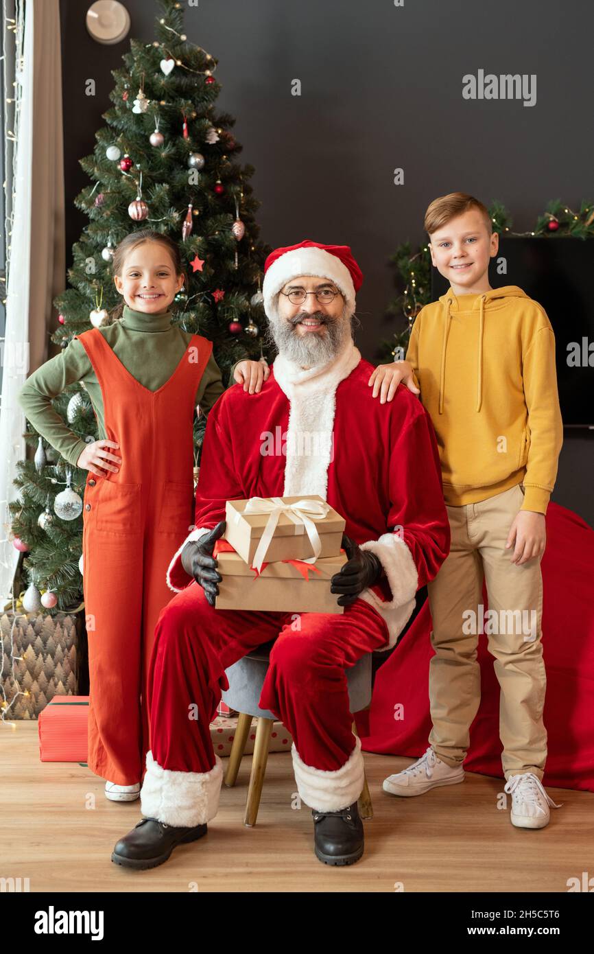 Porträt des lächelnden Weihnachtsmannes in rotem Kostüm sitzend mit Geschenken gegen den Weihnachtsbaum, während er mit Kindern posiert Stockfoto