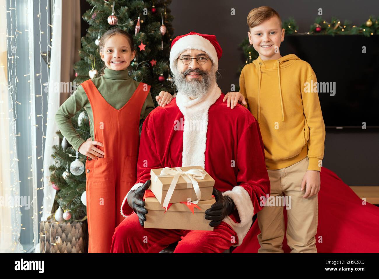 Portrait von lächelnden kaukasischen Kindern in hellen Outfits, die am Weihnachtsmann mit Weihnachtsgeschenken gegen geschmückten Baum im Wohnzimmer stehen Stockfoto