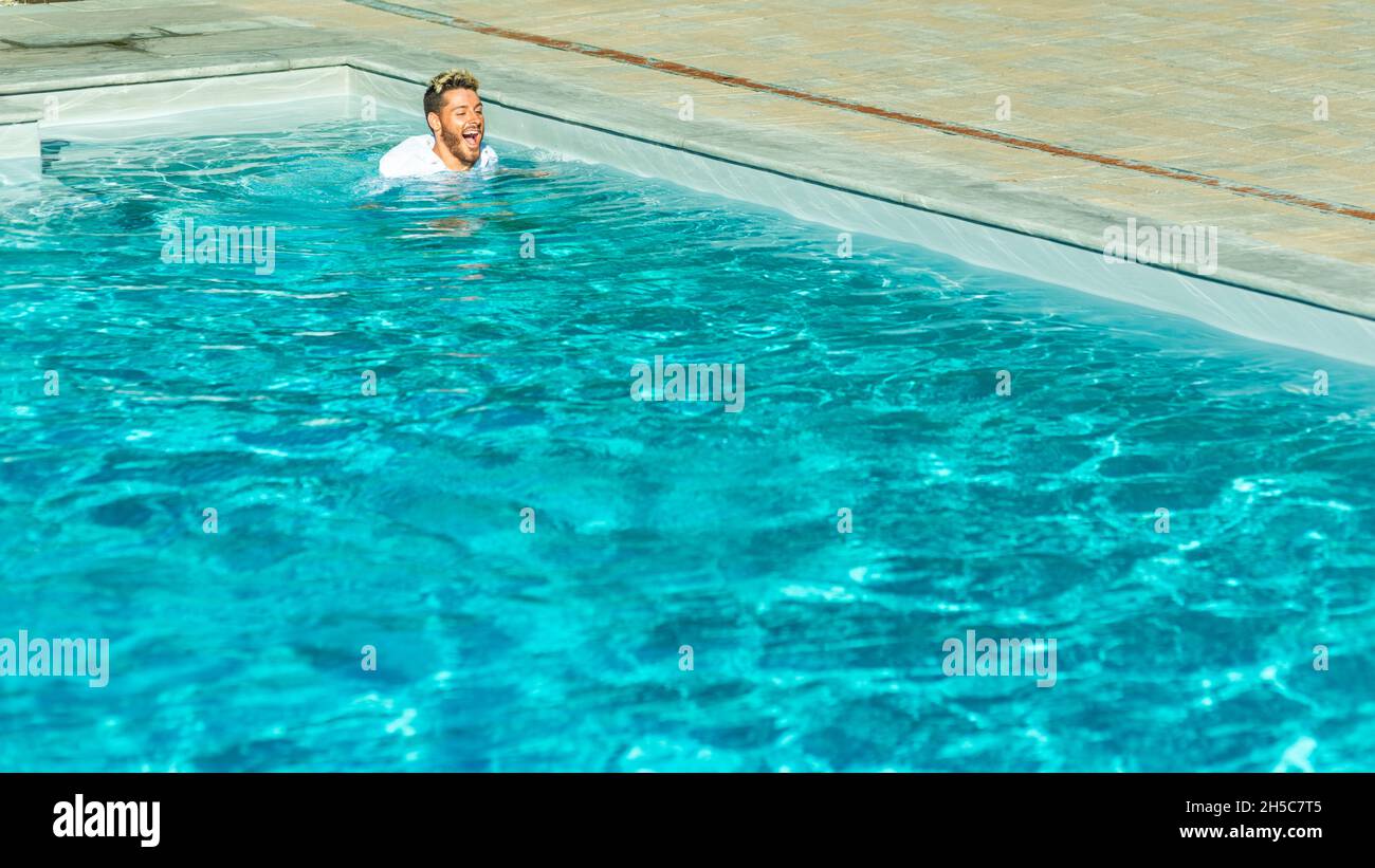 Fröhlicher junger Mann mit weißem Hemd, der in einem Pool mit kristallklarem, azurblauem Wasser schwimmt Stockfoto