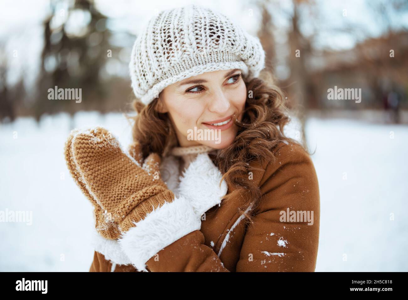 Lächelnde stilvolle Frau mittleren Alters mit Fäustlingen in einem Strickhut und Schaffell Mantel draußen im Stadtpark im Winter. Stockfoto