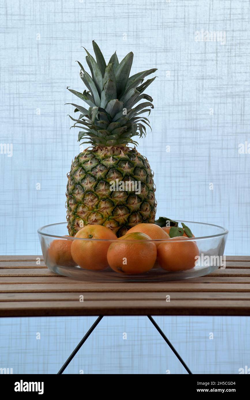 Glasschale mit Ananas und Orangen, auf einem Holzlattentisch und einem weißen strukturierten Hintergrund Stockfoto
