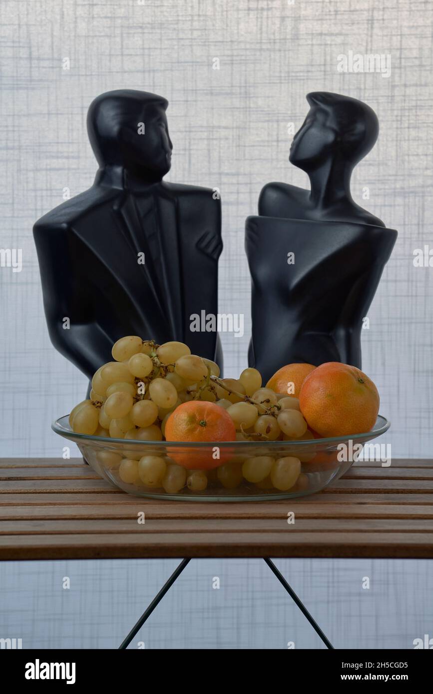 Glasplatte mit Trauben, Orangen und den schwarzen Skulpturen eines Mannes und einer Frau, auf einem Holzlattentisch und einem weißen strukturierten Hintergrund Stockfoto
