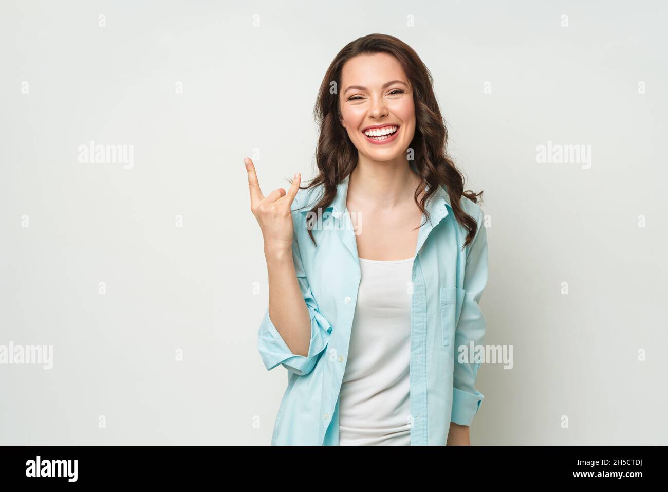 Emotive Brünette Frau machen Rock n Roll Zeichen, schreit laut, steht über weißem Hintergrund mit leerem Raum, fühlt sich selbstbewusst. Stockfoto