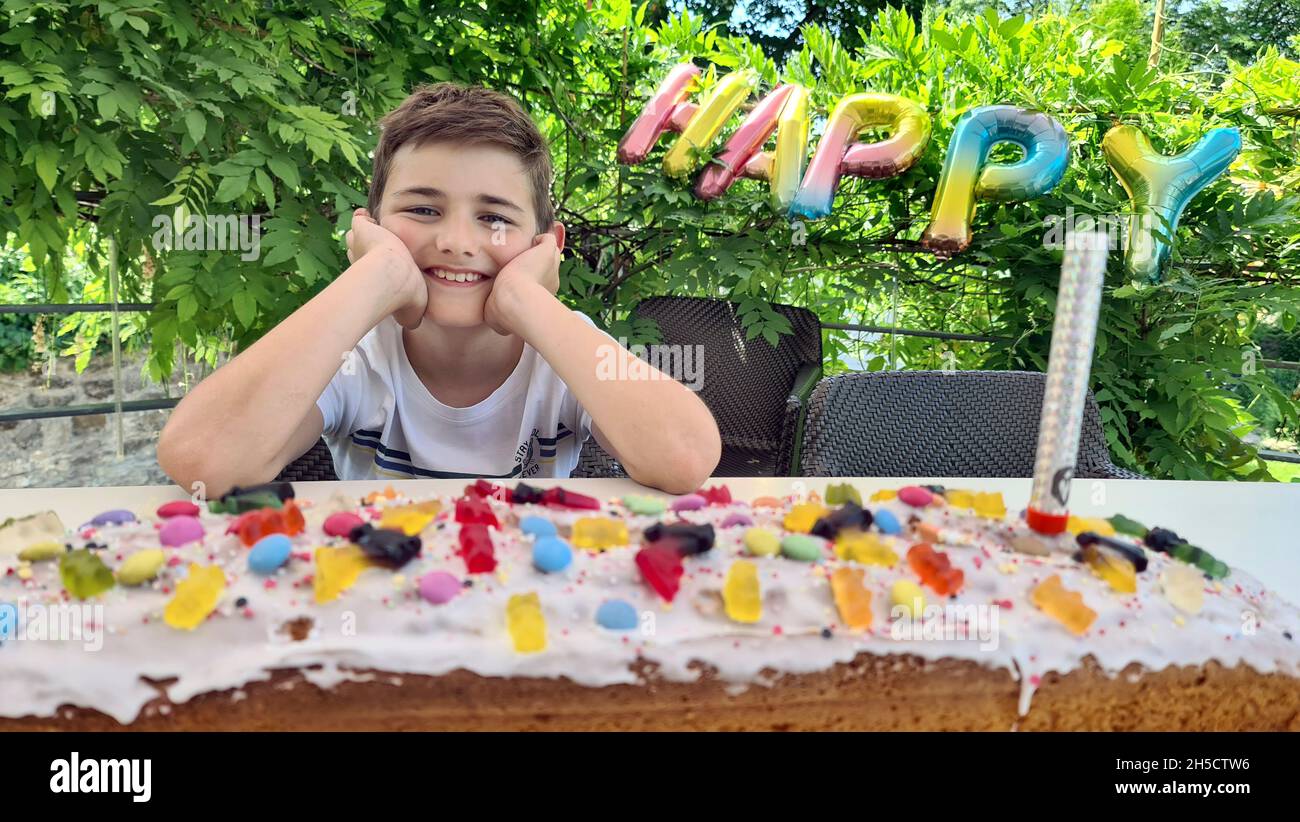 Happy Birthday Boy sitzt strahlend an einem Tisch mit einem riesigen Geburtstagskuchen Stockfoto