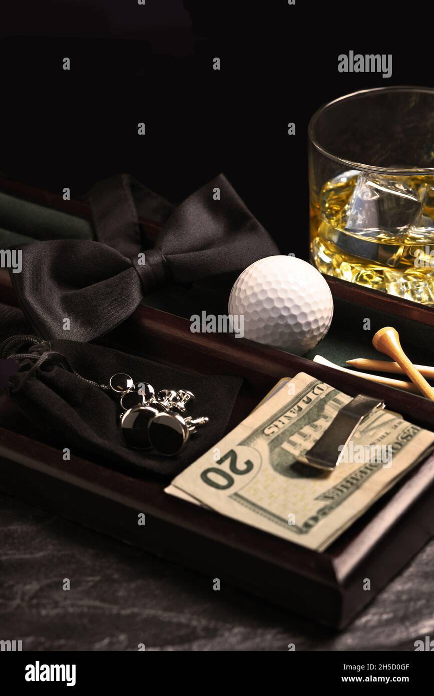 Nahaufnahme des Bureau Organizer für Herren. Im Tablett befinden sich Golfball-T-Shirts, Geldklammer, Fliege, Manschettenknöpfe und ein Glas Whiskey. Warmes Seitenlicht und Schalung Stockfoto