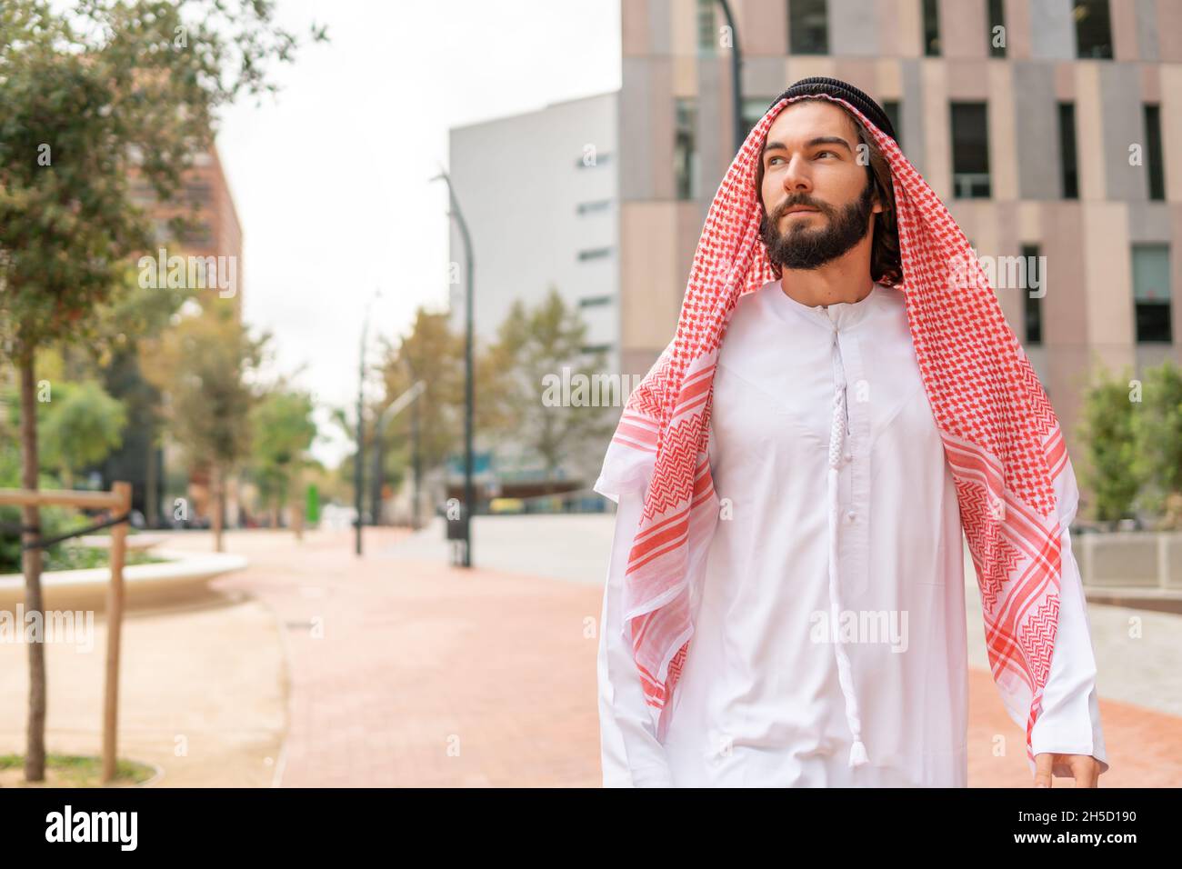 Bärtiger arabischer Mann in traditioneller Kleidung, der beim Spazierengehen im Park in der Innenstadt wegschaut Stockfoto