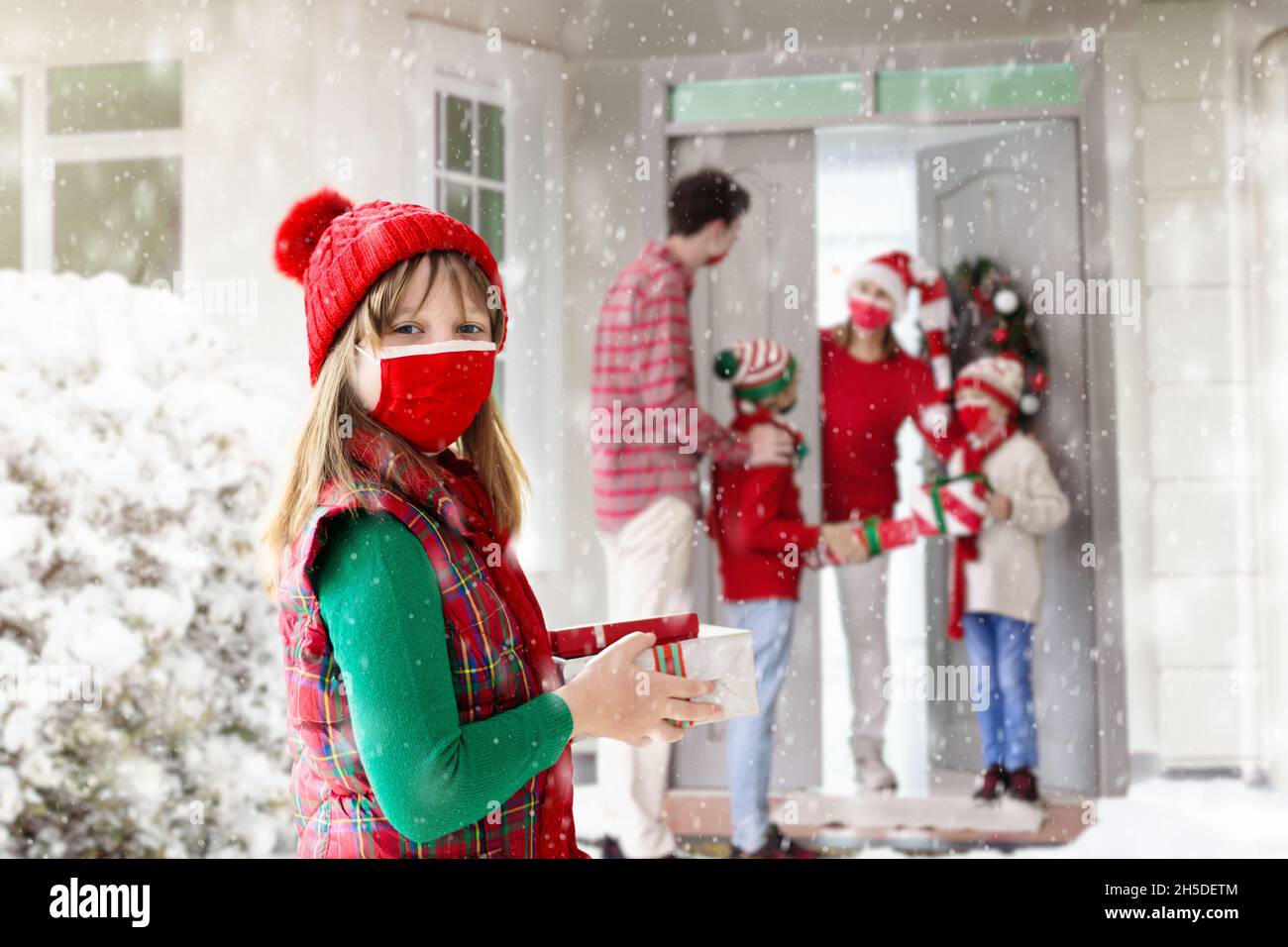 Kind in Gesichtsmaske mit Weihnachtsgeschenk an dekorierte Haustüre. Kind trägt Schutzmaske für sichere Weihnachtsfeier. Familienbesuch im Covid-19 Stockfoto