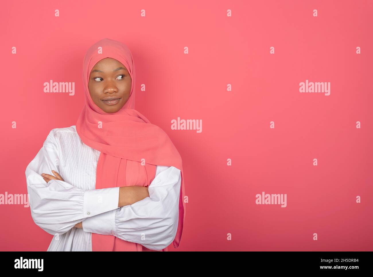 Zufriedene religiöse weibliche Modell hat die Arme gefaltet, sieht zur Seite, trägt rosa Kopftuch stehen auf rosa Hintergrund, kopieren Platz für Text Stockfoto