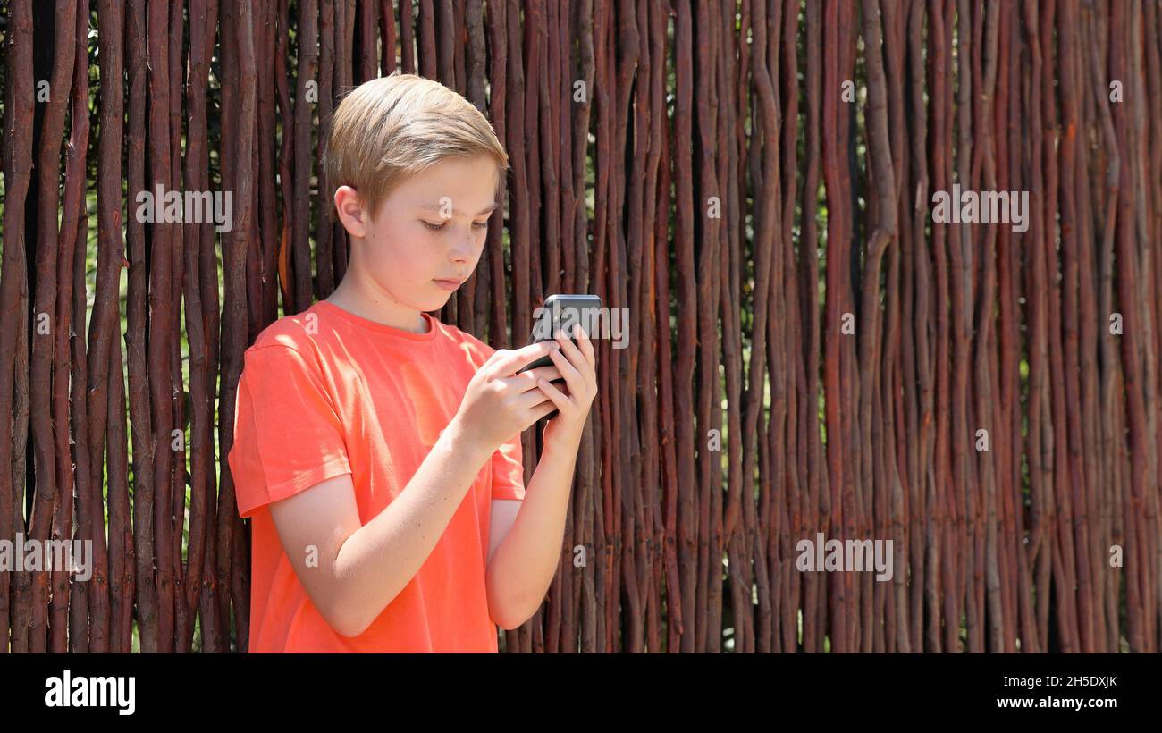 Porträt eines blonden Jungen mit orangefarbenem T-Shirt, der auf der Straße gegen einen Holzzaun steht und sein Handy benutzt Stockfoto