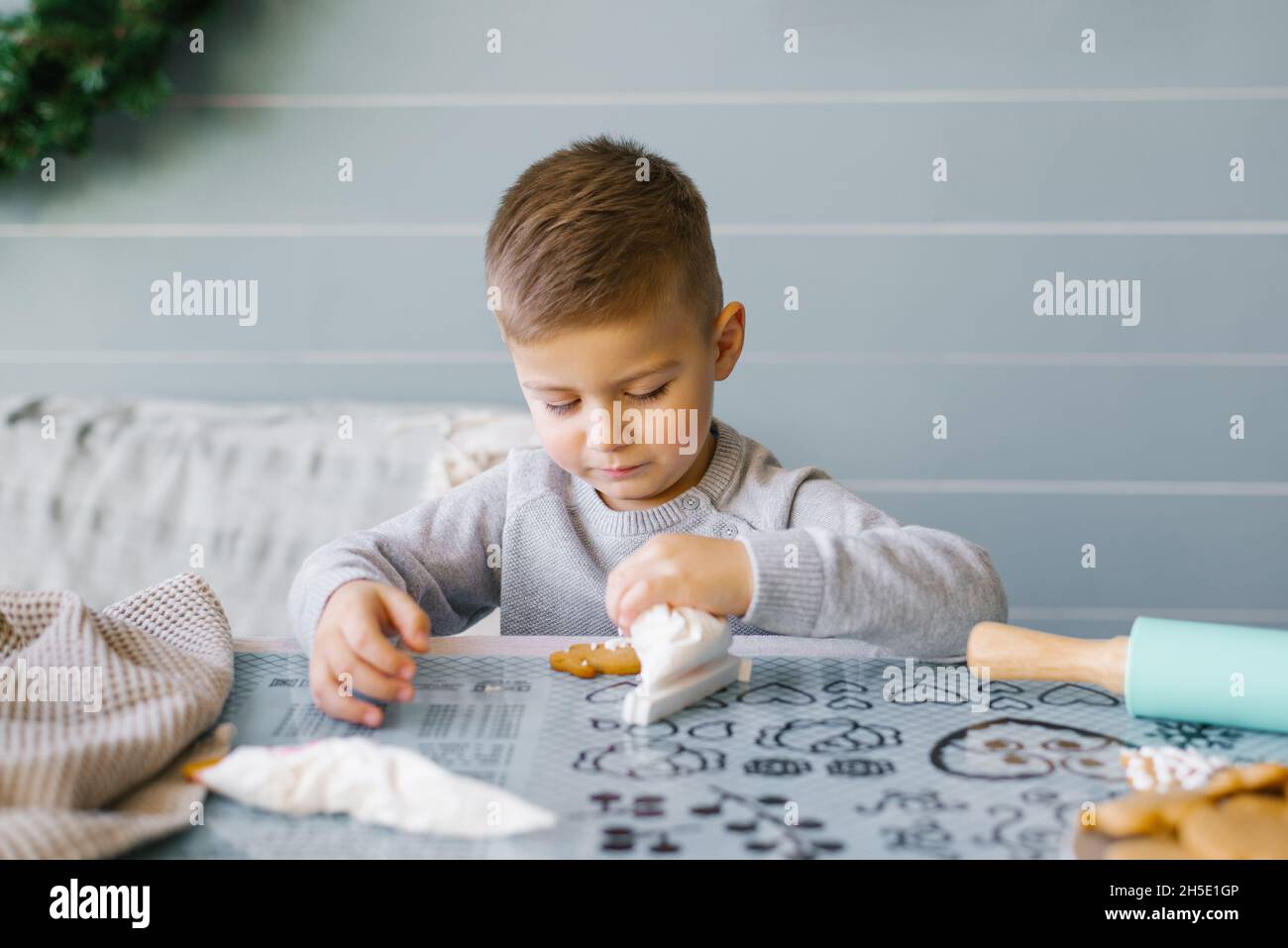 Ein schöner Junge sitzt auf dem Küchentisch und hilft seiner Mutter, Kekse zu backen und zu dekorieren Stockfoto