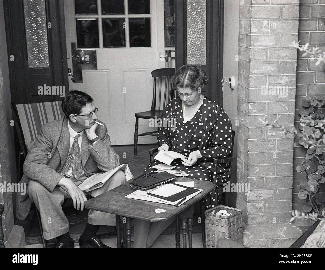 1937, ein historisches Paar, das auf der Veranda vor der Haustür sitzt, mit der Dame an einem kleinen Holztisch, die Papiere in ihrem Portemonnaie aussortierte und dem Herrn auf einem Liegestuhl mit einer Zeitung und einer Zigarette rauchte, England, Großbritannien. Sie tragen die formellen Kleider der Ära, die Dame in einem gepunkteten Kleid und den Herrn in Anzug und Krawatte. Stockfoto
