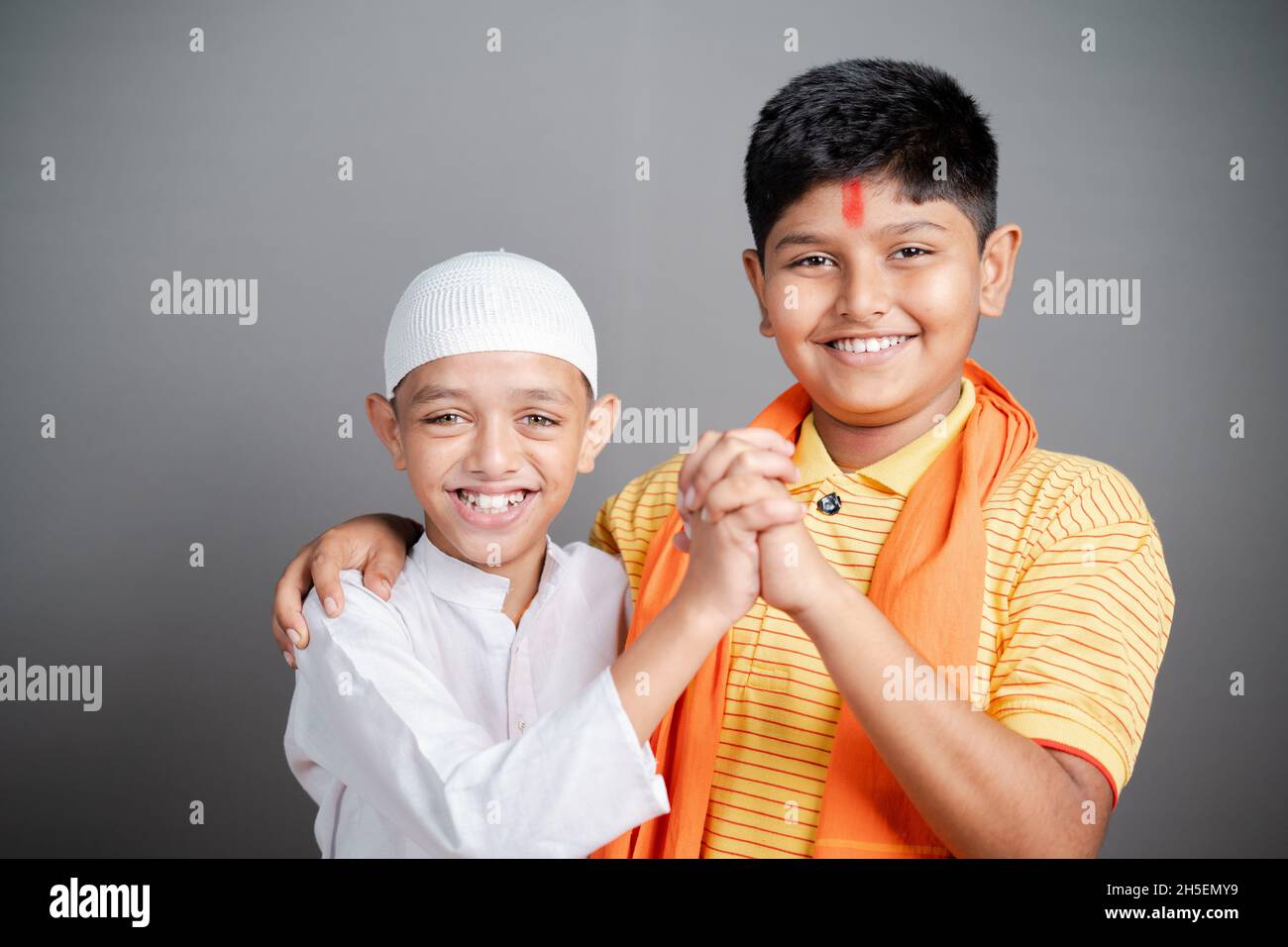 Glückliche Hindu-muslimische Kinder zeigen Einheit, indem sie die Hände zusammenhalten, indem sie die Kamera auf grauem Hintergrund betrachten - Konzept der Vielfalt, Bindung und Stockfoto