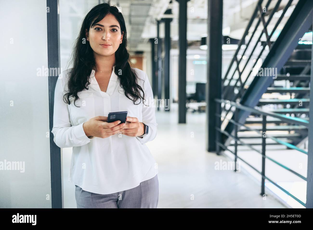 Eine intelligente Geschäftsfrau, die ein Smartphone in einem Büro hält. Selbstbewusste Geschäftsfrau, die auf die Kamera schaut, während sie allein an einem modernen Arbeitsplatz steht. Mittel-A Stockfoto