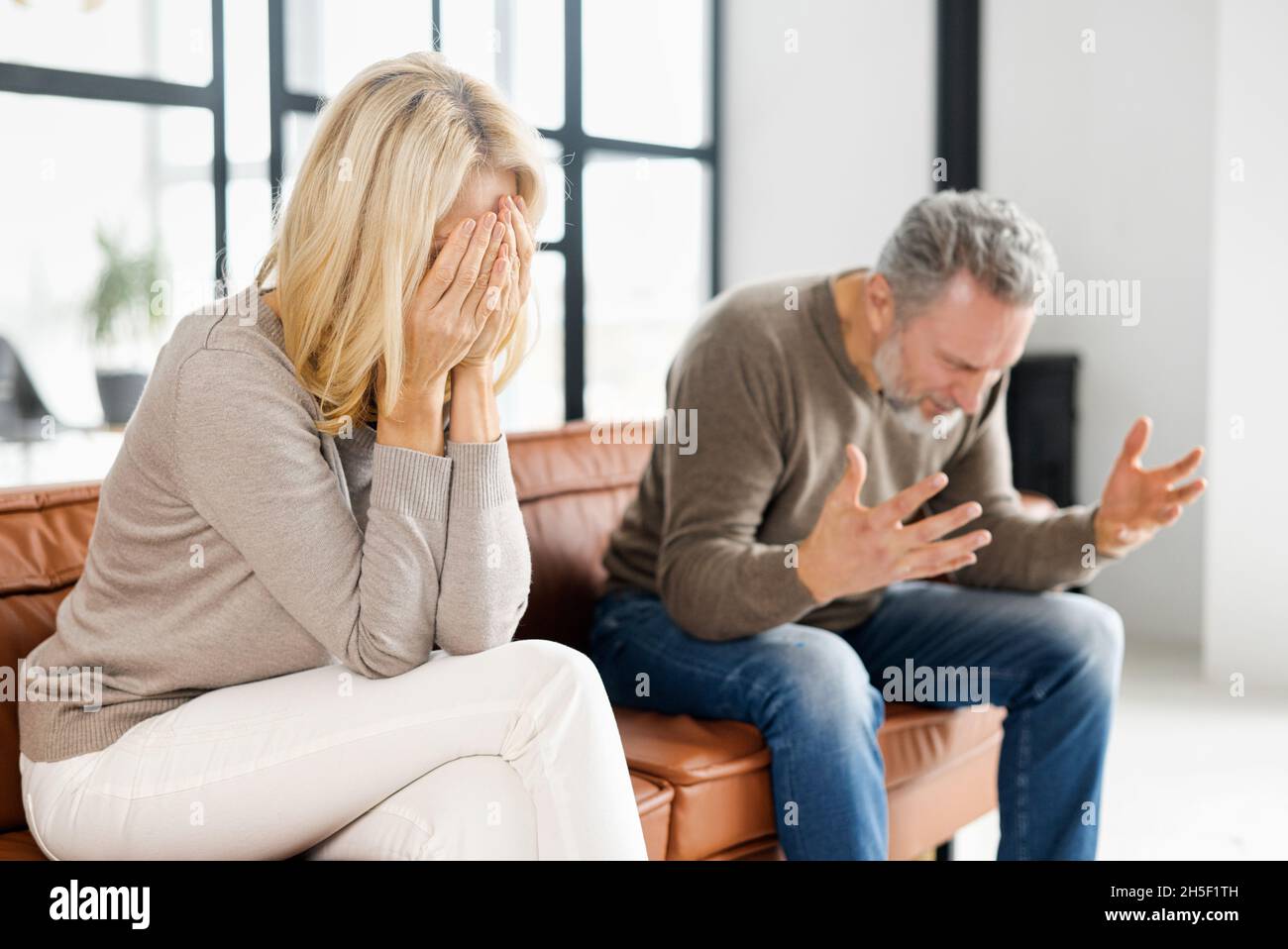 Hausmissbrauch-Konzept. Deprimierte Frau mittleren Alters sitzt auf der Couch und weint, ein gereizter und wütender reifer Mann schreit auf dem Hintergrund. Ein Paar streitet zu Hause Stockfoto