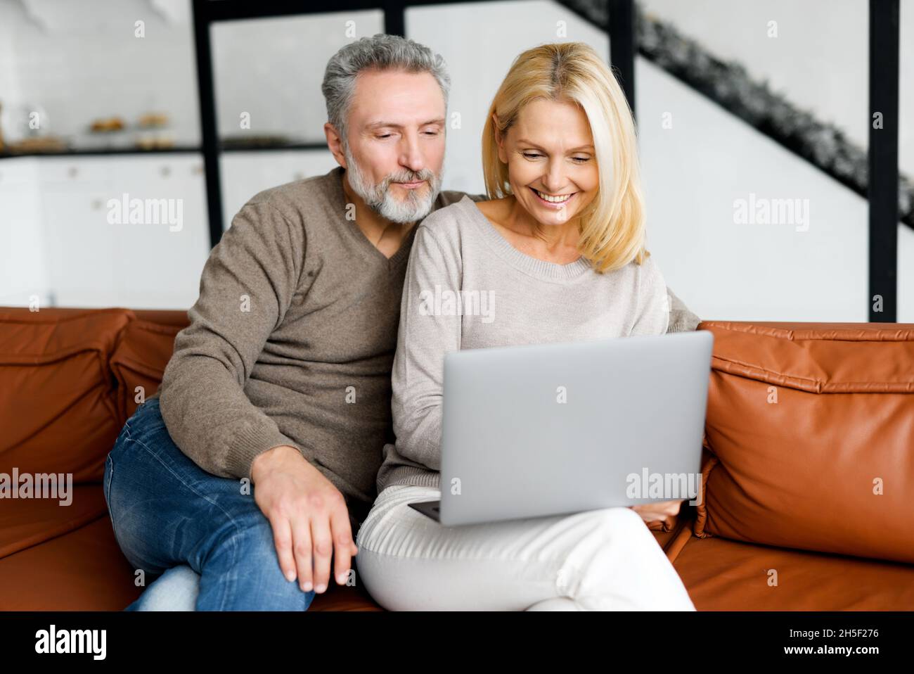 Paare mittleren Alters verbringen Freizeit mit einem Laptop zu Hause. Ehepartner sitzen auf der Couch, schauen sich Videos an, surfen im Internet oder kaufen online zusammen und genießen Zeit online Stockfoto