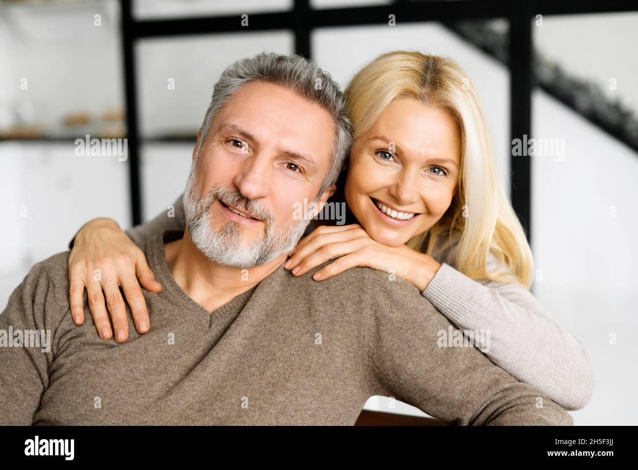 Liebe und Unterstützung Konzept. Porträt von Ehegatten mittleren Alters, glückliche, schöne Frau umarmt ihren grauhaarigen Mann, der auf der Couch sitzt, sie schauen auf die Kamera und lächeln Stockfoto