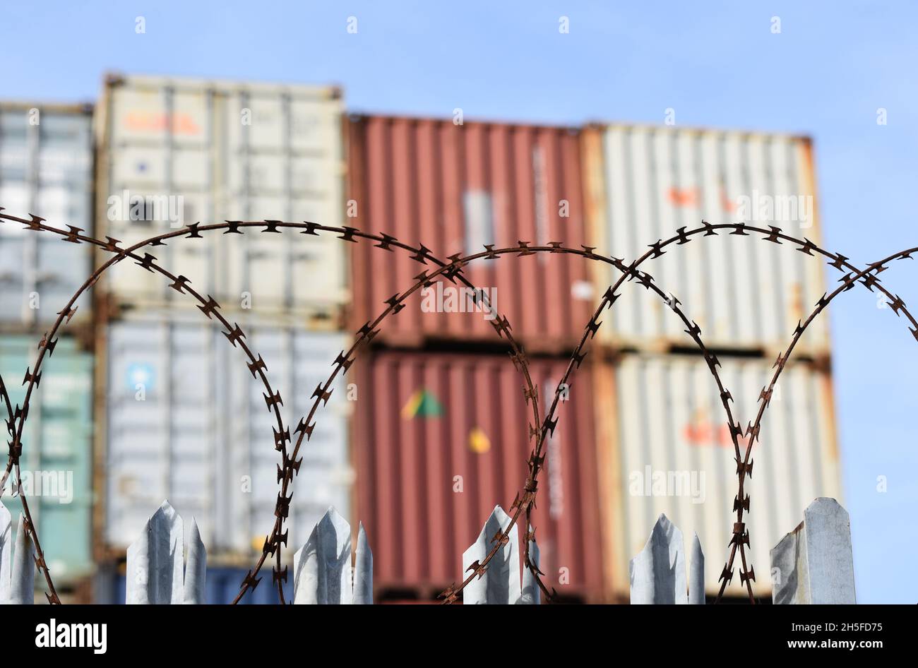 Frachtschiffcontainer, die hoch in einem geschäftigen britischen Hafen gestapelt sind, hinter einem Zaun mit Stacheldraht, der im Mittelpunkt steht. Stockfoto