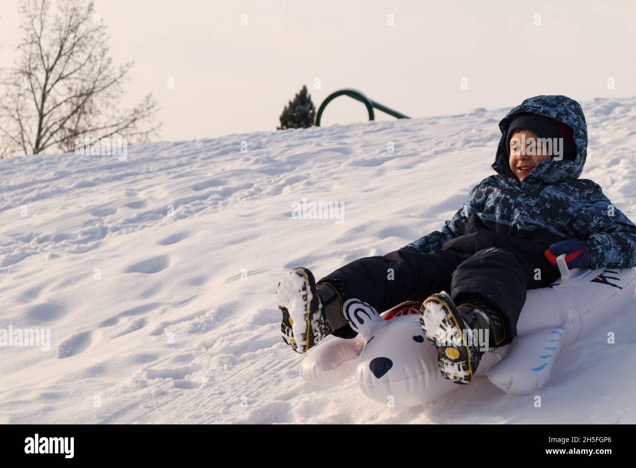 Kleiner Junge, der auf einer Rodel einen verschneiten Hügel hinunterrutscht Stockfoto