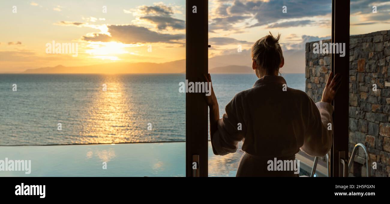 Frau im Bademantel, die zwischen Glasschiebetüren steht und den Sonnenaufgang über dem Meer in einer Luxusvilla genießt Stockfoto