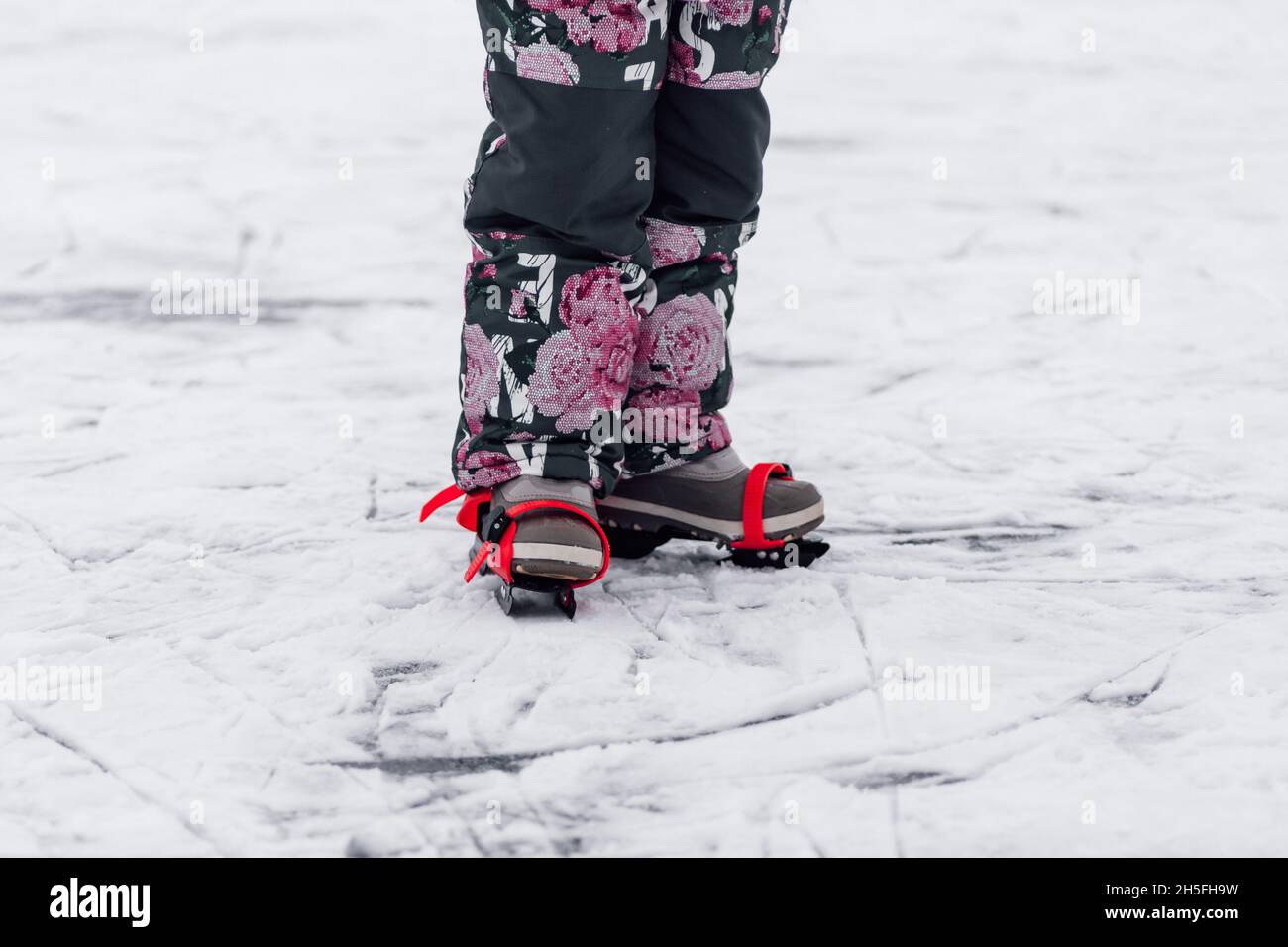 Nahaufnahme der Füße von Kindern. Kleines Mädchen in warmen Winteranzug steht auf Eis in Kinder Schlittschuhe mit roten Bändern an ihre Schuhe gebunden, verschneiten Winter Stockfoto