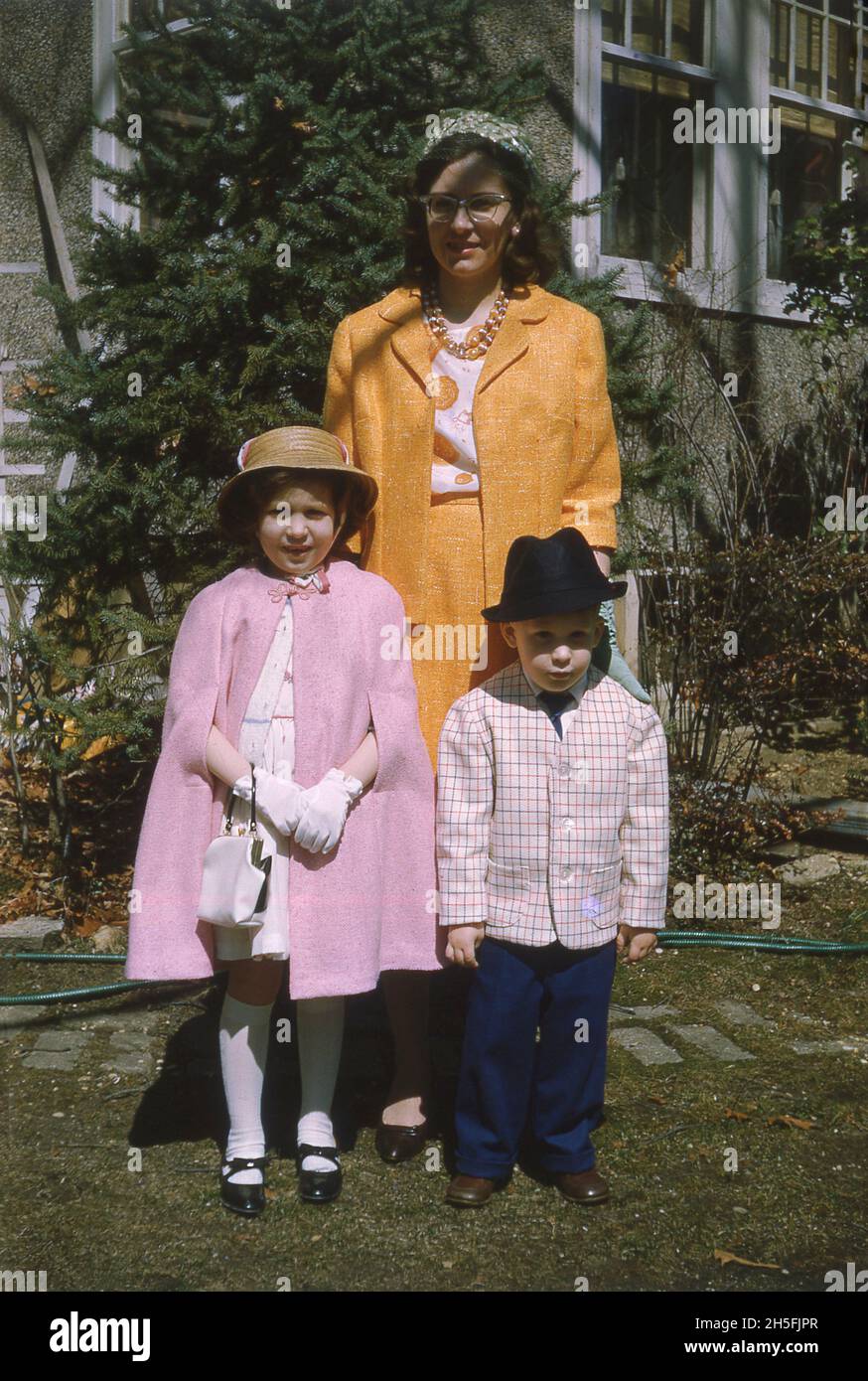 1967, März, Ostern, eine Mutter, die mit ihren zwei kleinen Kindern vor einem Haus steht, alle in Kleidung gekleidet, um an einem Osterdienst teilzunehmen, USA. Die Dame trägt einen orangen Ausstrichen, während das junge Mädchen in ganz rosa und auf ihrem Kopf eine Strohhaube trägt. Der kleine Junge sieht cool aus in einer hellrosa Jacke und einem schwarzen Hut. Stockfoto