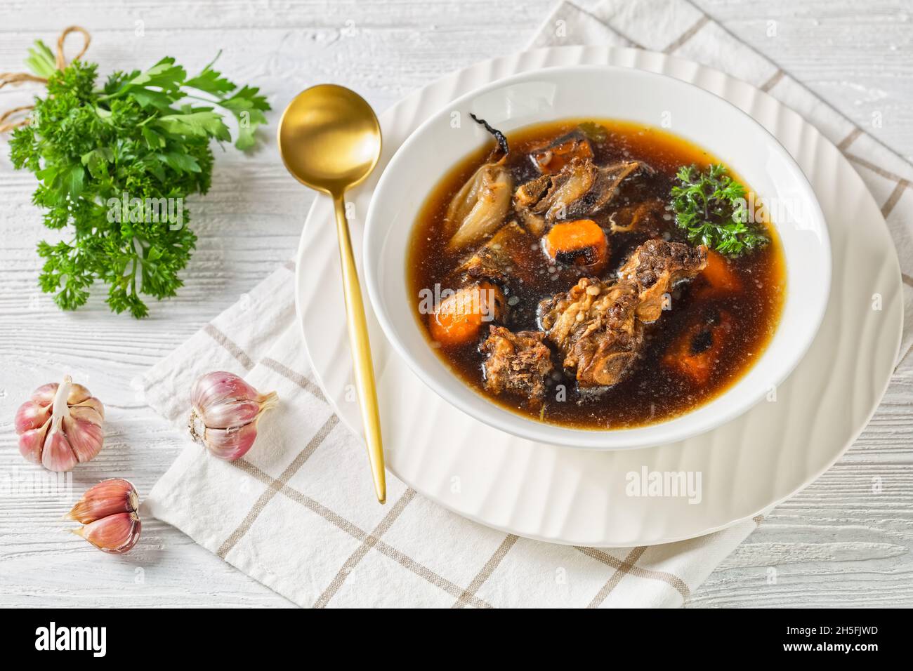 Rinderbrühe von Rinderfleisch auf Knochen langsam gekocht mit verkohltem Gemüse: Karotte, Zwiebel, Knoblauch und Gewürze in einer weißen Schüssel auf einem Holztisch serviert Stockfoto