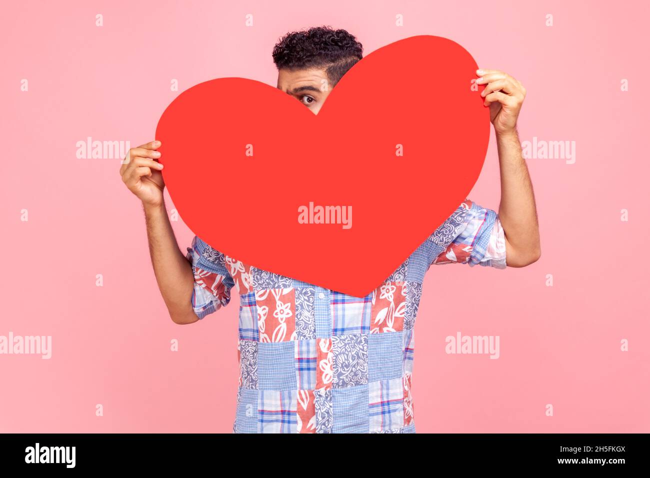 Positiver Mann mit dunklem Haar, der sich hinter einem großen roten Herzen versteckt und mit neugierigen neugierigen neugierigen Augen auf die Kamera blickt und Zuneigung liebt. Innenaufnahme des Studios isoliert auf rosa Hintergrund. Stockfoto