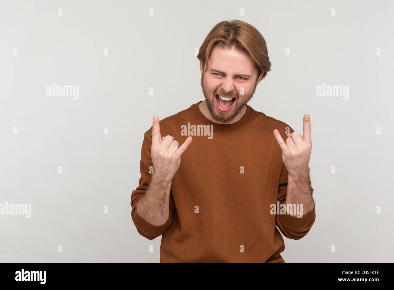 Porträt eines Mannes mit Bart, der Sweatshirt trägt, mit Rock and Roll-Zeichen-Handbewegung, schreiend, mit aufgeregt Gesichtsausdruck. Innenaufnahme des Studios isoliert auf grauem Hintergrund. Stockfoto