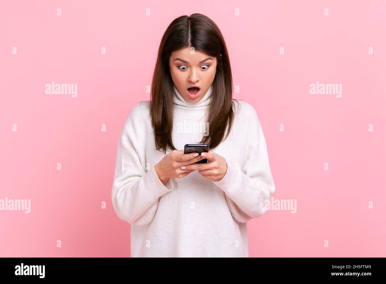 Porträt der schockierten schönen Bloggerin stehend, verwenden Smartphone mit staunenden Gesicht, geöffneten Mund, trägt weißen lässigen Stil Pullover. Innenaufnahme des Studios isoliert auf rosa Hintergrund. Stockfoto
