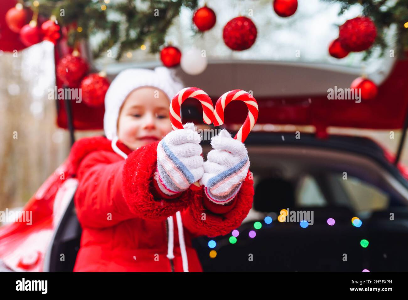 Blonde Mädchen in roter Jacke und weißen Hut haben Spaß mit roten und weißen süßen Sticks in geschmückten weihnachtsauto Stockfoto