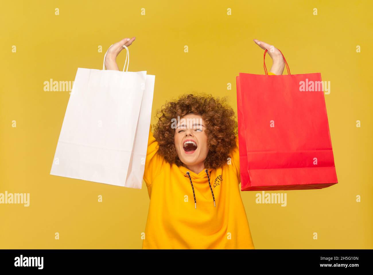 Porträt einer aufgeregten überglückten Frau mit Afro-Frisur, erhobenen Armen mit Einkaufstaschen, fröhlich brüllend, mit lässigem Hoodie. Innenaufnahme des Studios isoliert auf gelbem Hintergrund. Stockfoto
