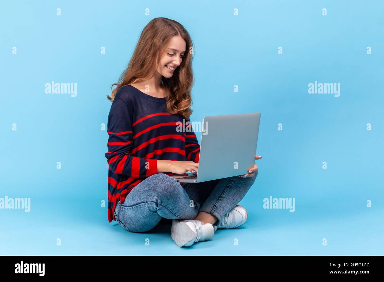 Die positive Frau trägt einen gestreiften Pullover im lässigen Stil, sitzt mit gekreuzten Beinen und arbeitet online auf dem Schoß und ist mit ihrem freiberuflichen Job zufrieden. Innenaufnahme des Studios isoliert auf blauem Hintergrund. Stockfoto