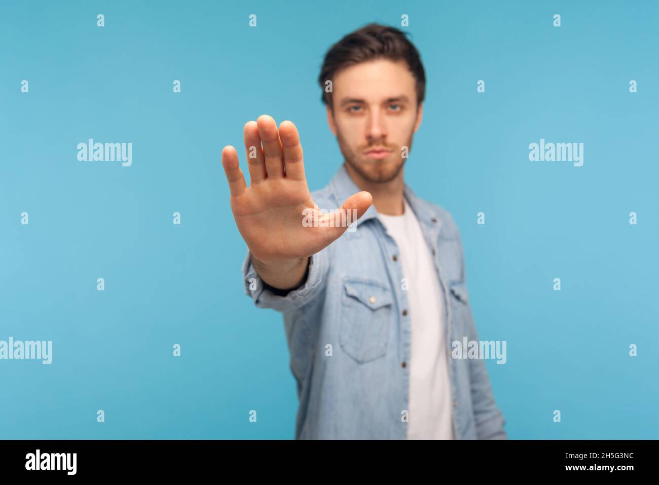 Porträt eines wütenden oder aggressiven Mannes, der Denim-Hemd trägt, mit Stopphand steht und mit strengem und ernsthafteren Ausdruck auf die Kamera blickt. Innenaufnahme des Studios isoliert auf blauem Hintergrund. Stockfoto