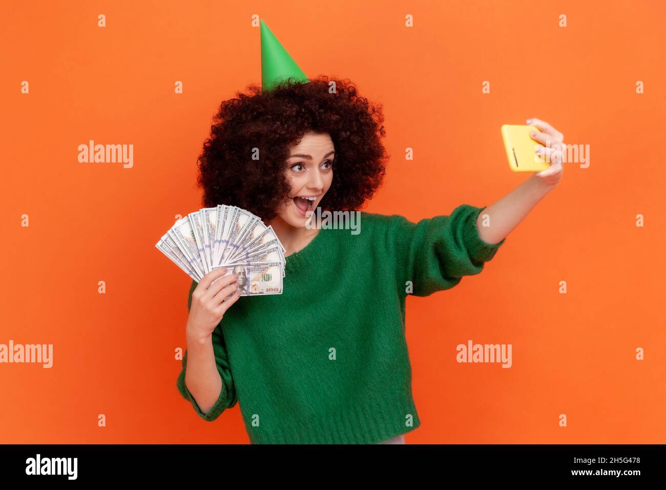 Zufriedene, aufgeregt Frau mit Afro-Frisur, die einen grünen Pullover im lässigen Stil trägt und einen Partykegel mit Dollarschein hält und Livestream sendet. Innenaufnahme des Studios isoliert auf orangefarbenem Hintergrund. Stockfoto