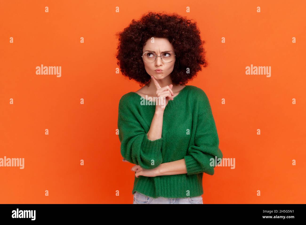 Nachdenkliche Frau mit Afro-Frisur trägt grünen Pullover im Casual-Stil und Brillen, die denken, Kinn halten, wegschauen, tief in Gedanken sind. Innenaufnahme des Studios isoliert auf orangefarbenem Hintergrund. Stockfoto