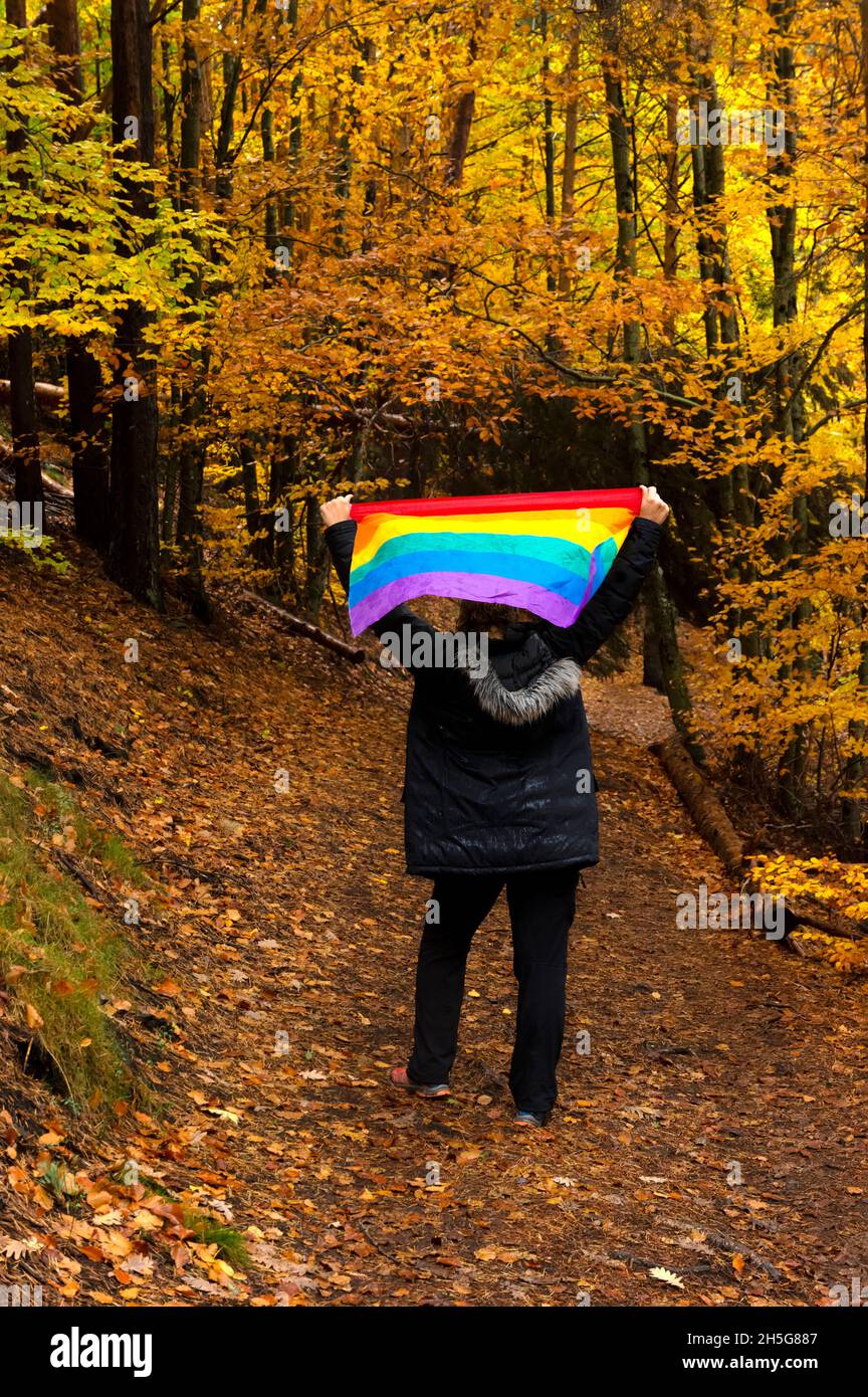 Nicht erkennbare Frau auf dem Rücken mit einem Mantel in einem tausendjährigen Buchenwald, der im Herbst im Regen eine Regenbogenfahne über ihren Kopf hebt. Stockfoto