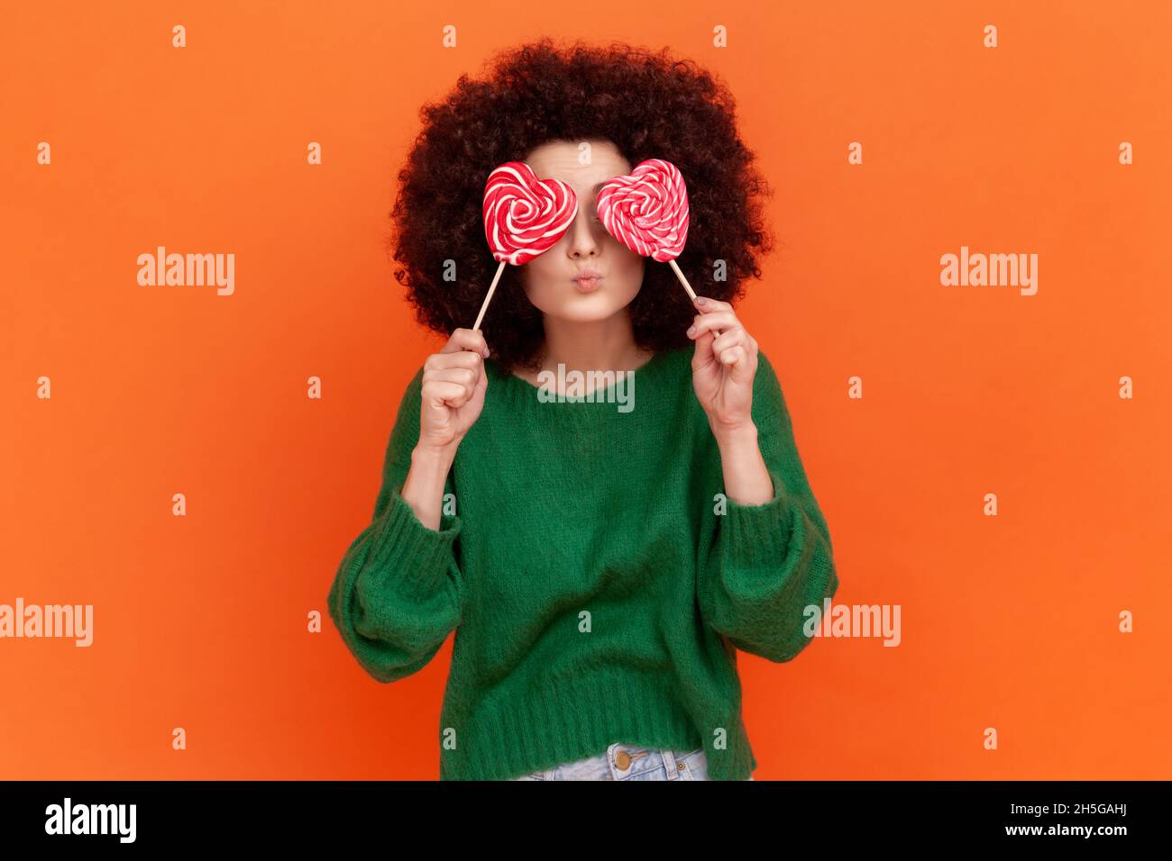 Romantische Frau mit Afro-Frisur trägt grünen lässigen Stil Pullover, die ihre Augen mit herzförmigen Süßigkeiten und Pout Lippen, Kuss Geste. Innenaufnahme des Studios isoliert auf orangefarbenem Hintergrund. Stockfoto