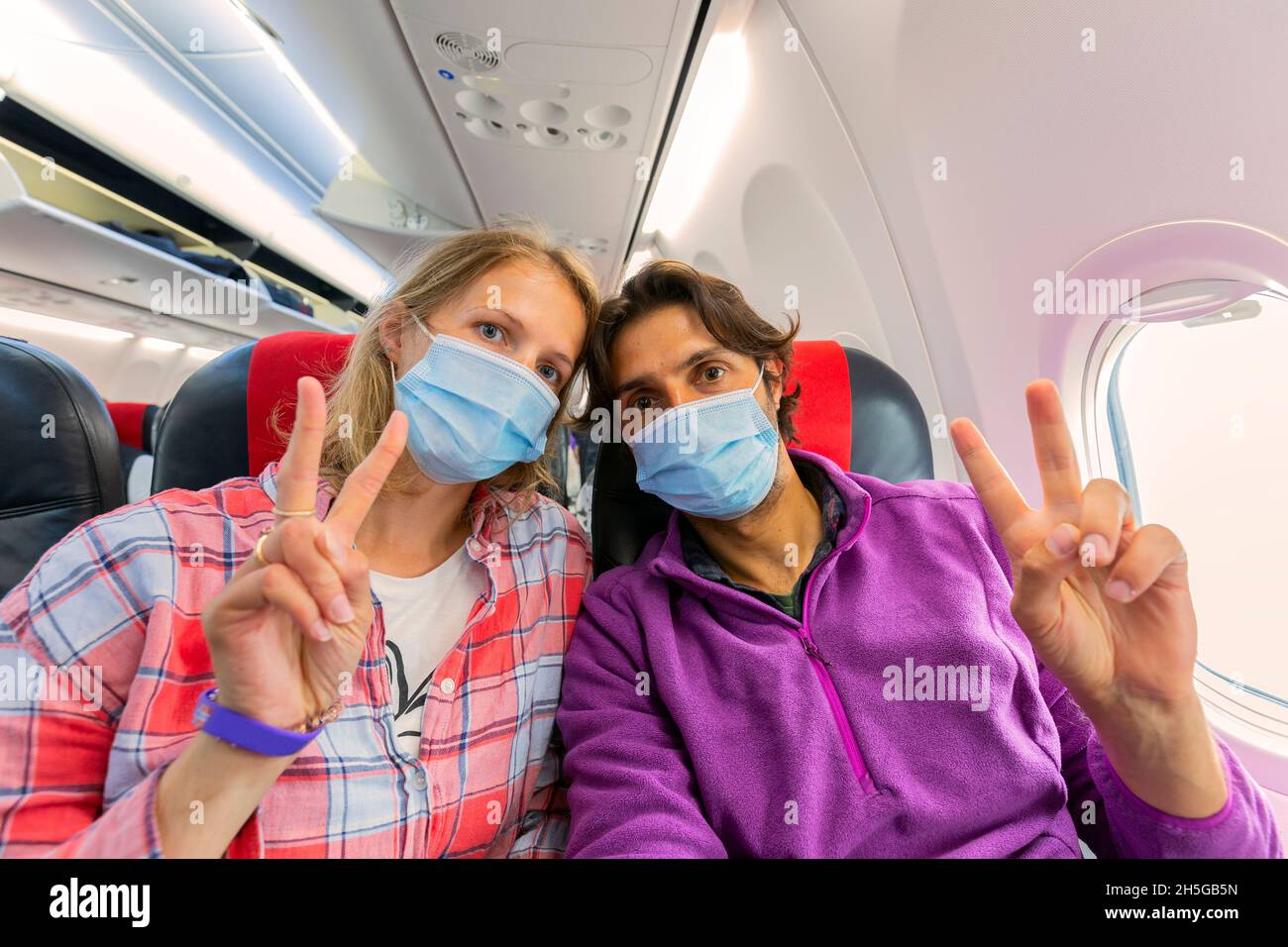 Das junge Paar trägt eine Gesichtsmaske für COVID-19-Schutz und sitzt auf einem Fenstersitz im Flugzeug, auf dem dem das V-förmige Schild mit zwei Fingern auf der linken Hand des Kamerasitzers zu sehen ist Stockfoto