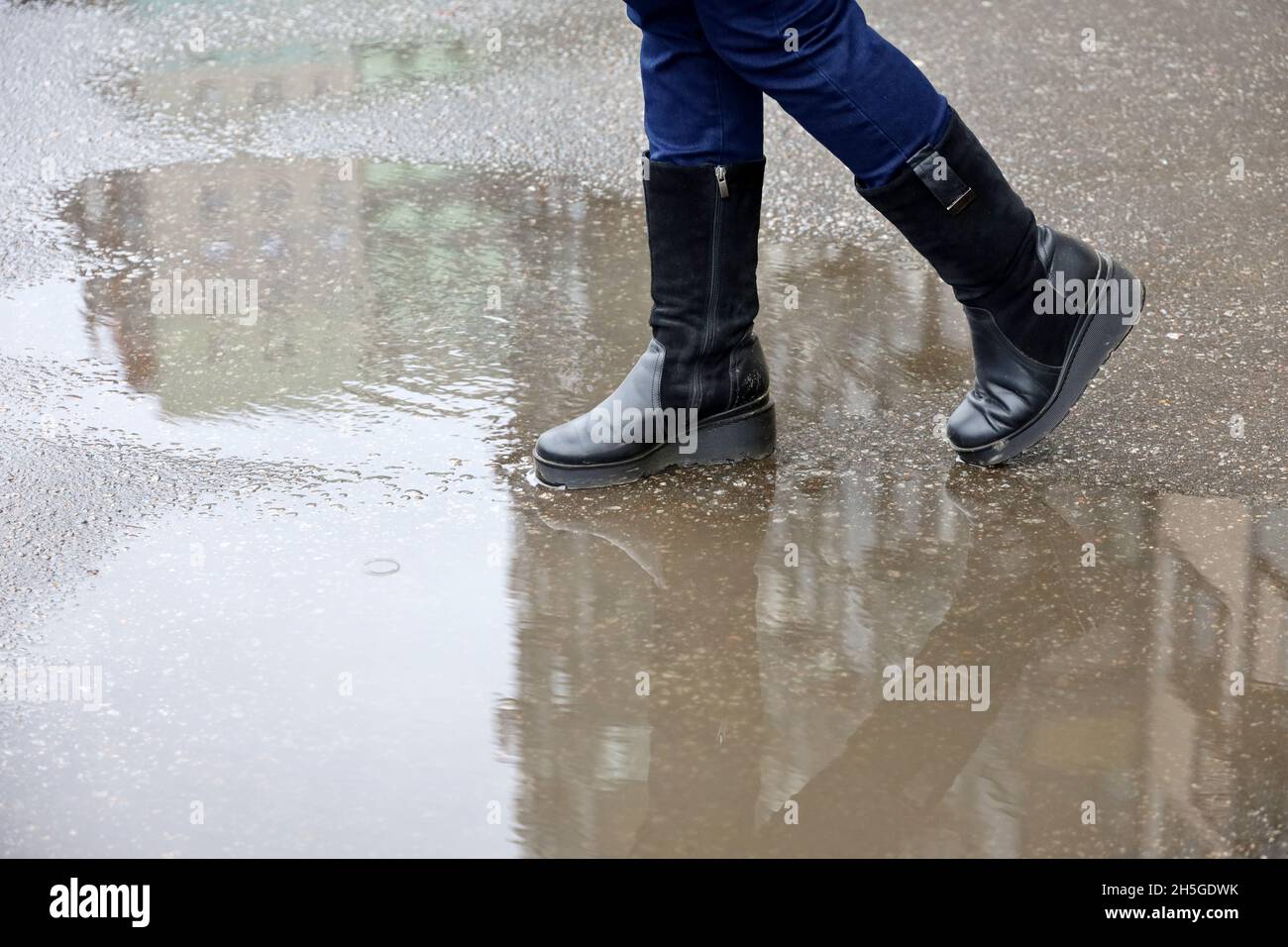 Regen in der Stadt, weibliche Beine in Stiefeln auf einer Straße mit Pfützen. Wohngebäude spiegeln sich im Wasser, Frau geht im Herbstwetter Stockfoto