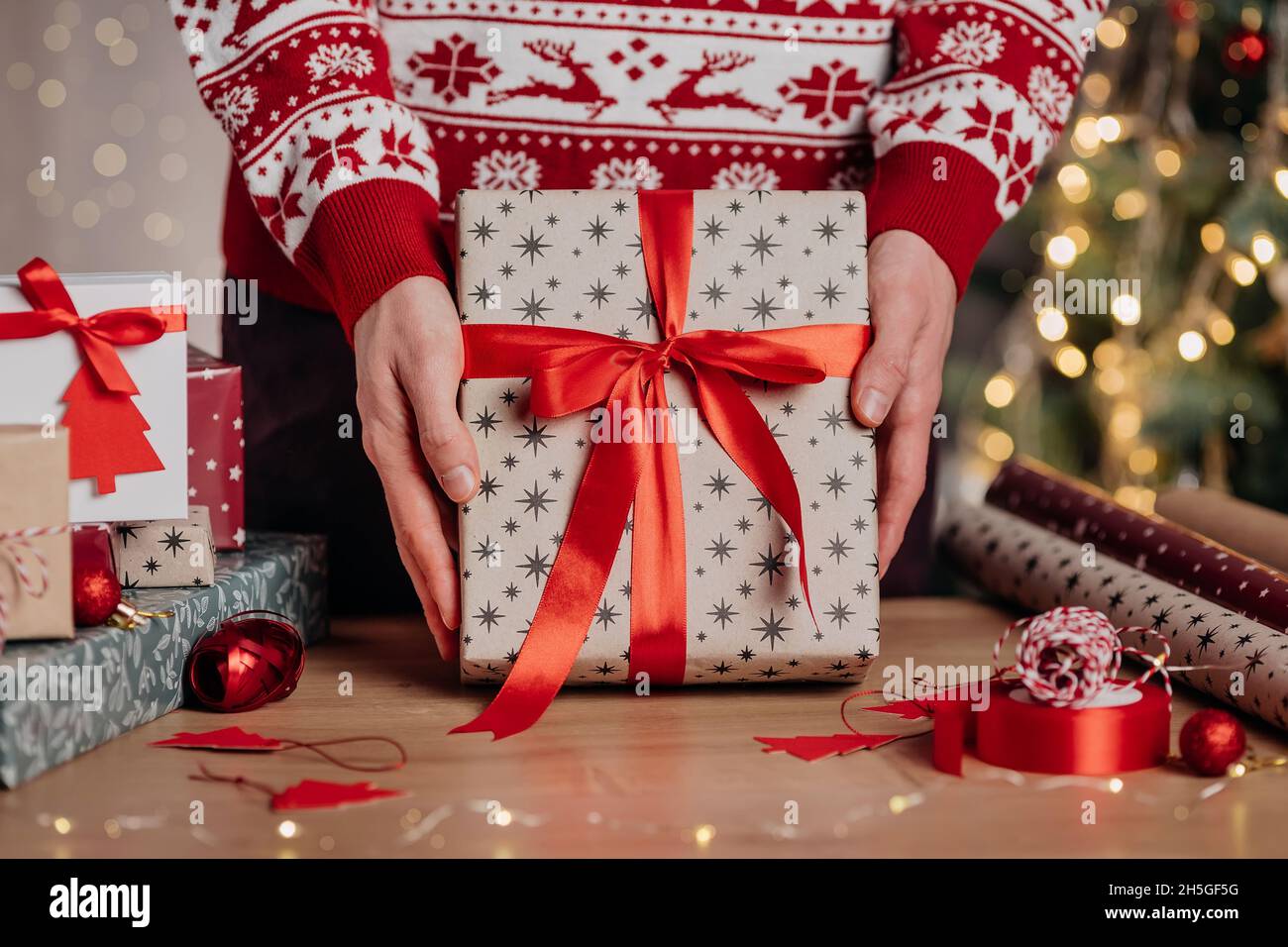 Mann hält in den Händen schöne Weihnachten Geschenkbox in Kraftpapier und rotem Band auf dem Hintergrund von weihnachtsbaum und Lichter verpackt. Präsentiert die Vorbereitung auf Stockfoto