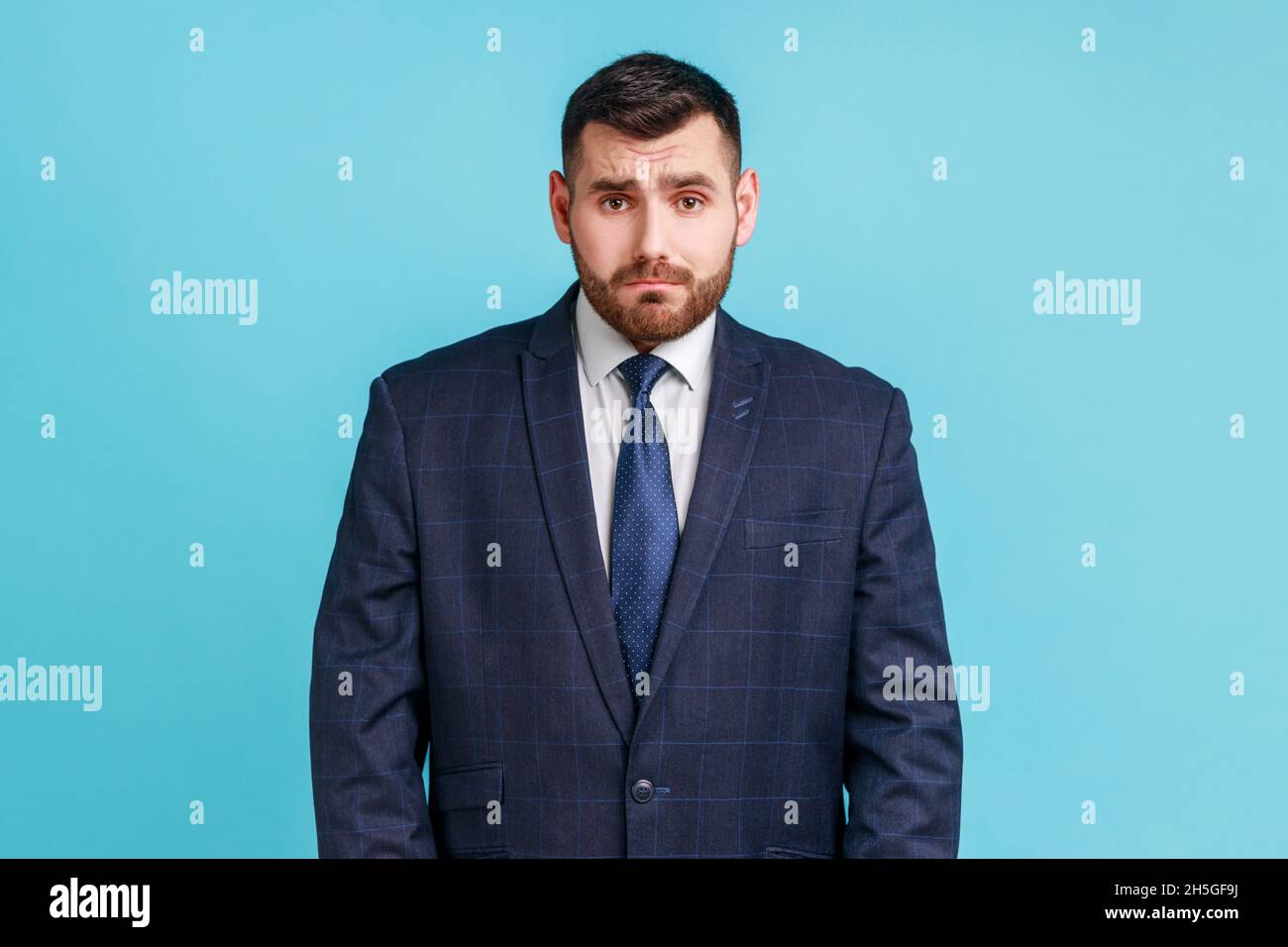 Attraktiver unglücklicher bärtiger Geschäftsmann, der einen dunklen Anzug im offiziellen Stil trägt und mit trauriger Gesichtsausdruck auf die Kamera blickt und Probleme hat. Innenaufnahme des Studios isoliert auf blauem Hintergrund. Stockfoto