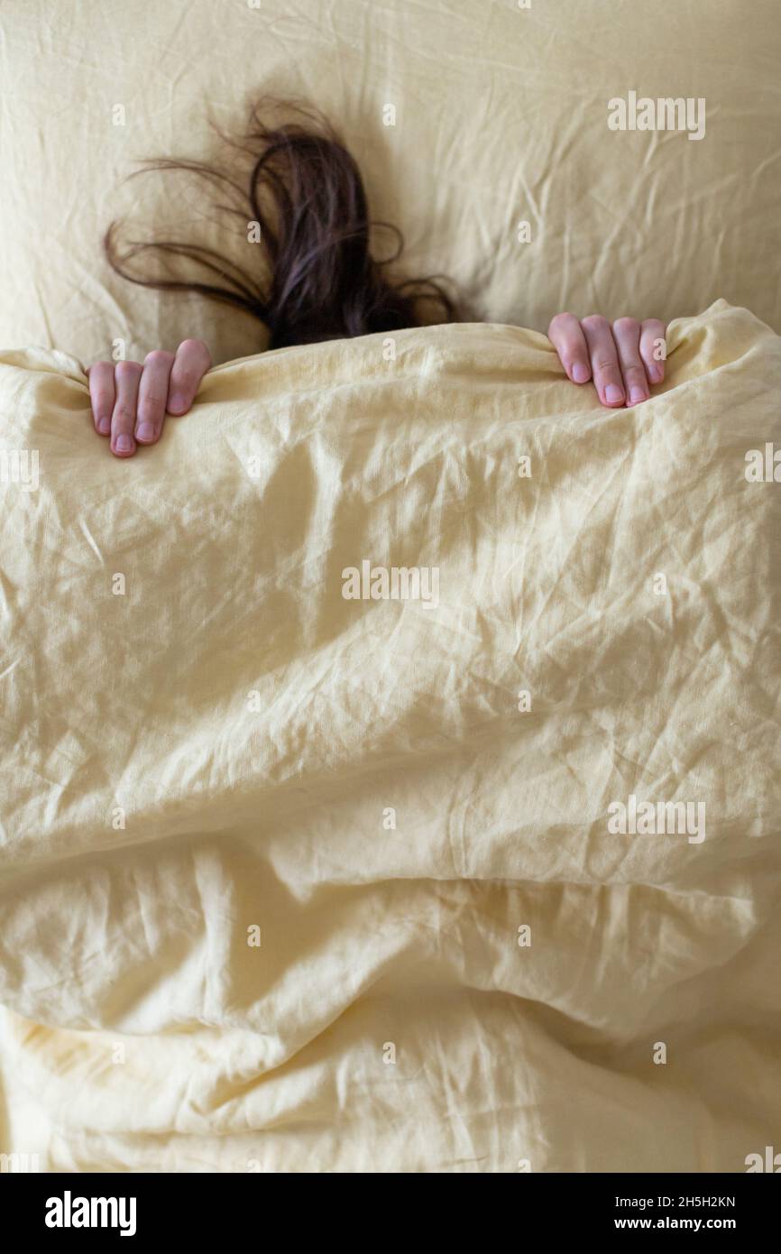 Das Mädchen im Bett ist mit einer gelben Decke mit dem Kopf bedeckt, man sieht einen Teil ihrer Hände und Haare. Das Konzept, sich vor Herbst und Winter zu verstecken. Stockfoto