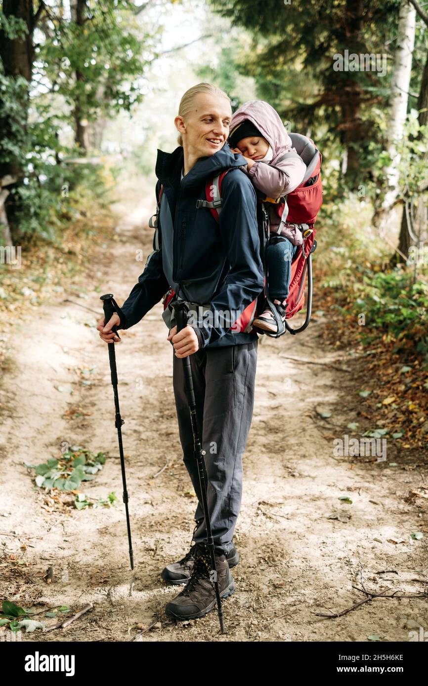 Mann, der mit einem verklenkten Kleinkind in den Berg wandert. Kind sitzt im Rucksack auf dem Rücken des Vaters. Aktives Familienabenteuer im Wald mit müdem Mädchen. Kalt Stockfoto