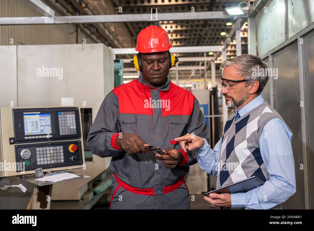 Multirassische industrielle Mitarbeiter stehen neben CNC-Steuerung und reden über den Produktionsprozess. Schwarze Arbeiter Messen Mit Messschieber. Stockfoto